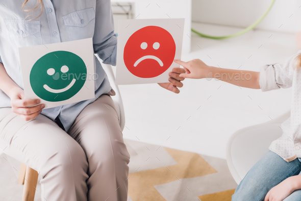 cropped shot of psychologist showing happy and sad emotion faces cards to child