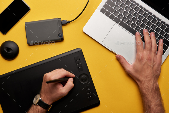 cropped shot of graphics designer working with laptop and graphics tablet on yellow surface