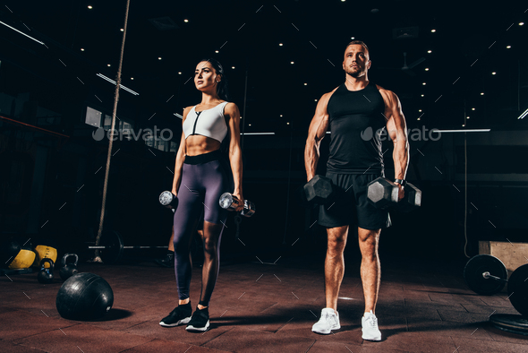 fit sportsman and sportswoman exercising with dumbbells together in dark gym