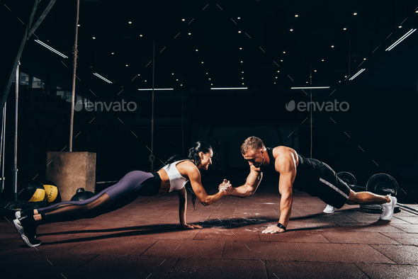 athletic sportsman and sportswoman doing push ups together and holding hands in dark gym