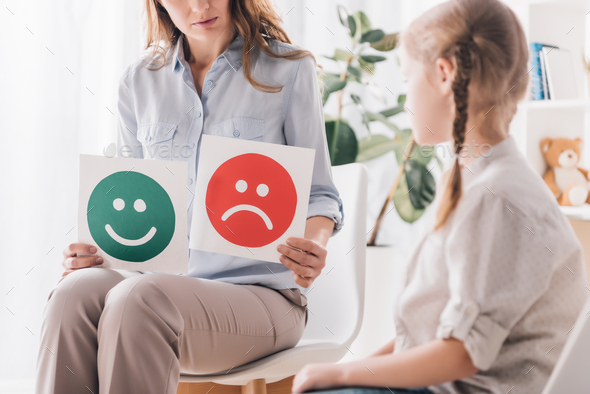 cropped shot of psychologist showing happy and sad emotion faces cards to child
