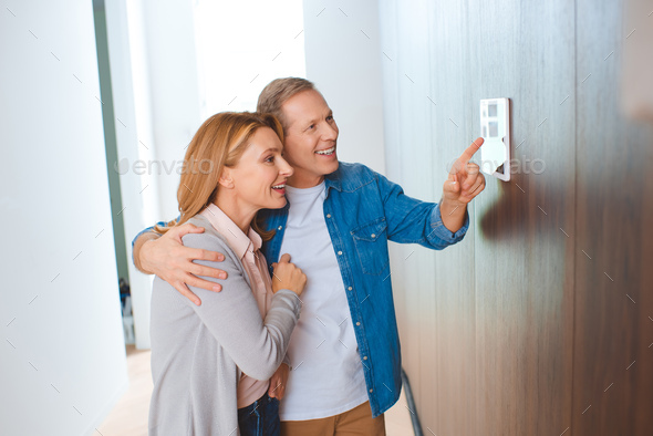 happy man pointing at smart home control panel while hugging wife