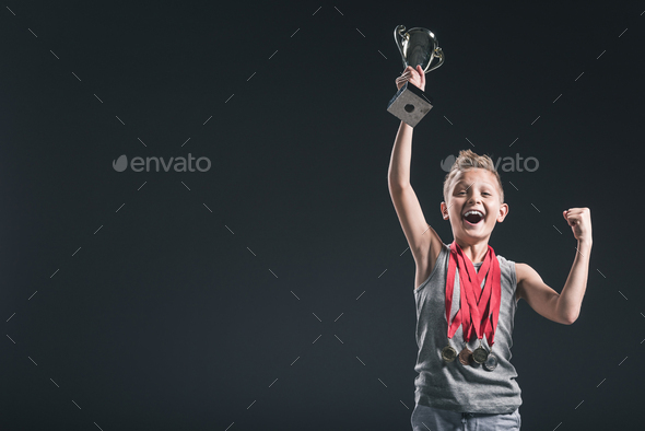 portrait of happy boy in sportswear with medals and champions cup gesturing on black background