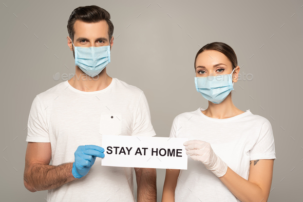 Young couple in medical masks holding card with stay at home lettering isolated on grey