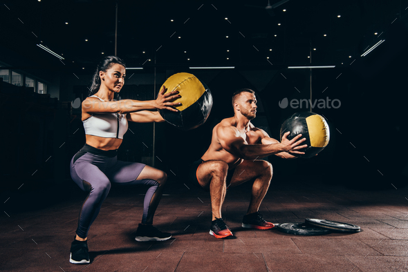 athletic sportsman and sportswoman doing squats with medicine balls together in dark gym