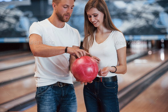 Hold it by that way. Man teaching girl how to holds ball and play bowling in the club