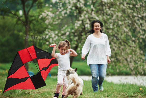 Positive female child and grandmother running with red and black colored kite in hands outdoors