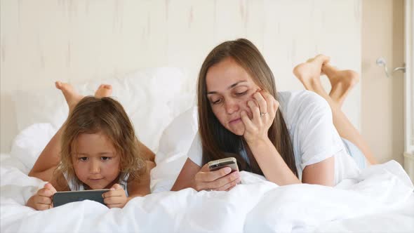 Young Mother with Little Cute Daughter are Using Gadgets Lying on a White Bed