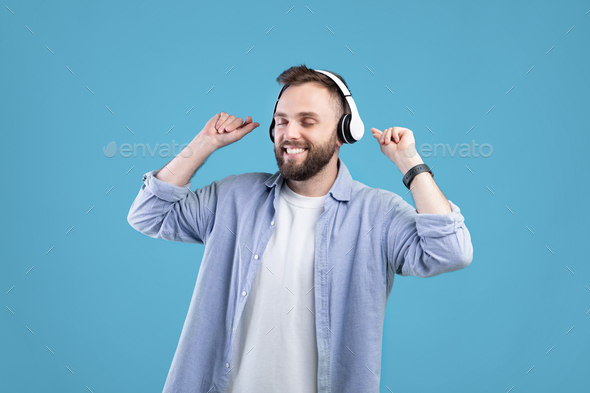 Happy young guy in headphones dancing to his favorite song, listening to music on blue studio