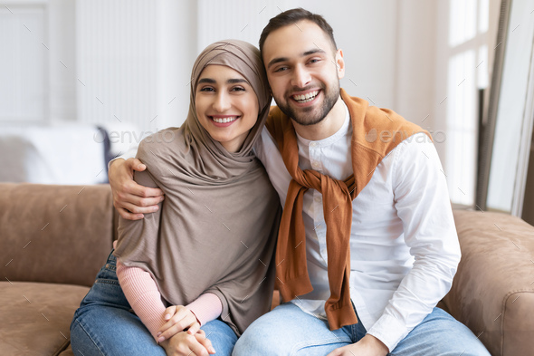 Happy Muslim Couple Embracing Sitting Together On Couch At Home Stock Photo  by Prostock-studio