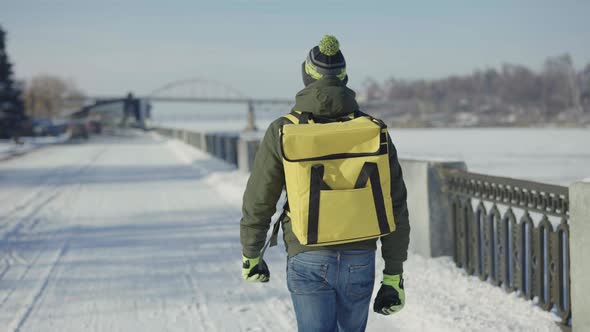 Back View of Courier with Large Yellow Thermo Bag Walking Along Snow Covered City Street alt