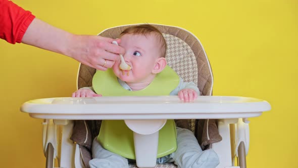 Mom feeding toddler baby from a spoon on a high chair for children, studio yellow background.