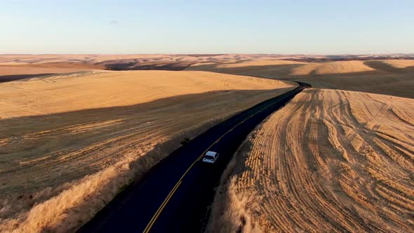 Drone follow shot of a car driving along a narrow country road among golden fields at sunrise or sun alt