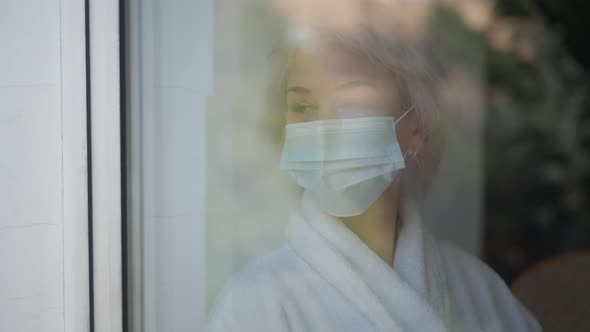 Shooting Through Window Sad Mature Woman in Covid19 Face Mask Standing Indoors Looking Out alt