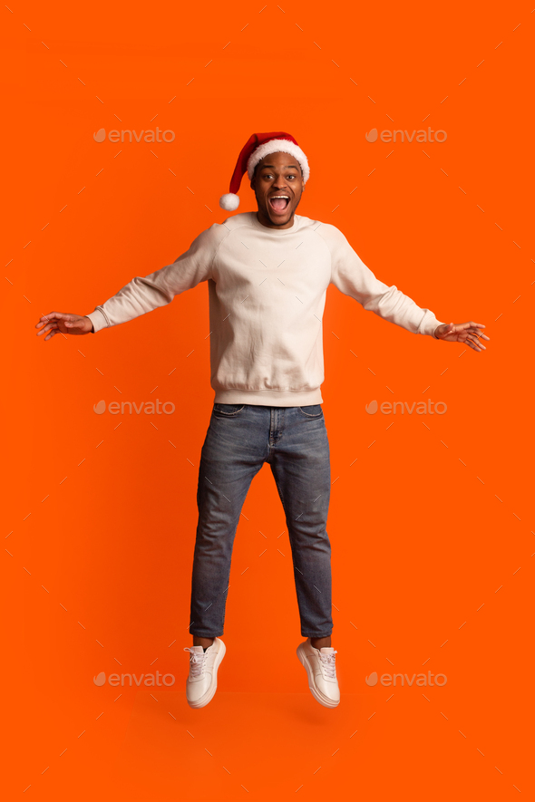 Emotional Surprised Black Guy In Santa Hat Jumping On Orange Background