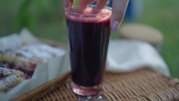 Female Hand Putting Black Currant Juice in Glass on Picnic Basket in Slow Motion alt