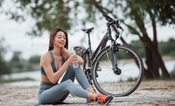 Drinking water. Female cyclist with good body shape sitting near her bike on beach at daytime