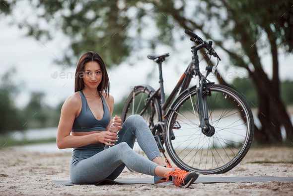 Human water balance. Female cyclist with good body shape sitting near her bike on beach at daytime