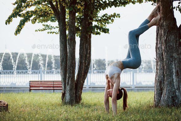 Using tree to keep balance. Young woman with slim type of body does exercises in the park