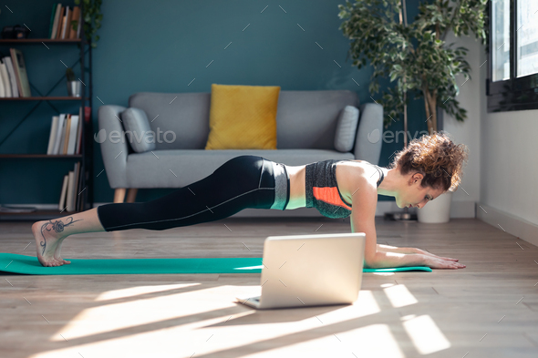 Sporty young woman doing hypopressive exercises following online gym classes via laptop at home.