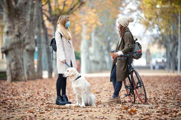 Two friends wearing surgical mask talking and walking with their dog and bike in the park in autumn.