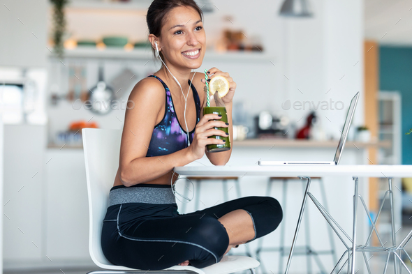 Sporty woman drinking detox juice while working with laptop after exercises in the kitchen at home. - Stock Photo - Images