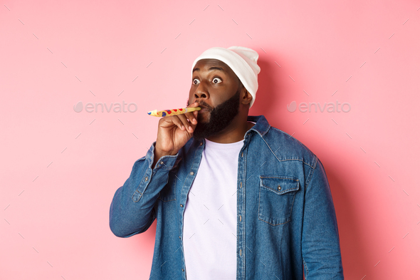 Funny Black man blowing in party whistle and pouting, staring sideways with popped eyes, standing