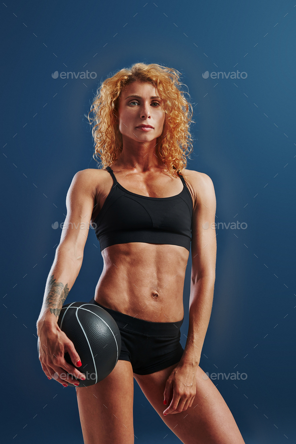 Stands with black soccer ball. Redhead female bodybuilder is in the studio on blue background