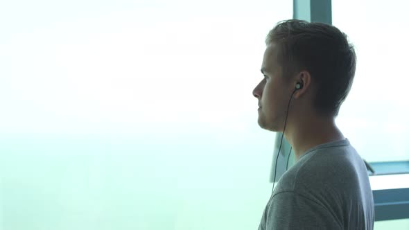 Young Man Stands Near a Big Window Listens to Music in Headphones