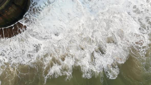 Aerial footage shot from overhead looking down at the sea crashing against a sea wall. alt