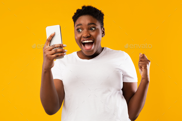 African Woman Holding Smartphone Shaking Fists In Joy, Yellow Background