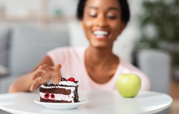Diet breakdown concept. Young black lady reaching for cake instead of apple, choosing junk food over