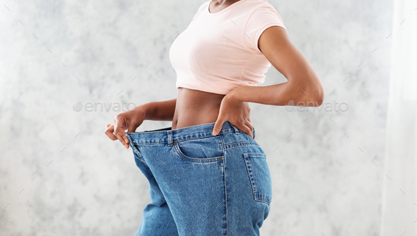 Unrecognizable black woman in oversized jeans showing results of her weight loss diet or liposuction