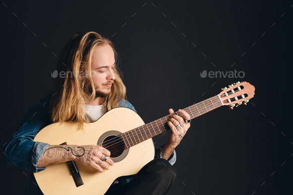 Portrait of tattooed musician with long hair playing acoustic guitar.  Stylish guy with guitar Stock Photo by garetsworkshop