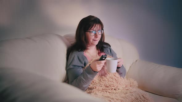 Happy Woman with a Glass of Coffee on the Couch at Night in Front of the TV Switches Channel an alt