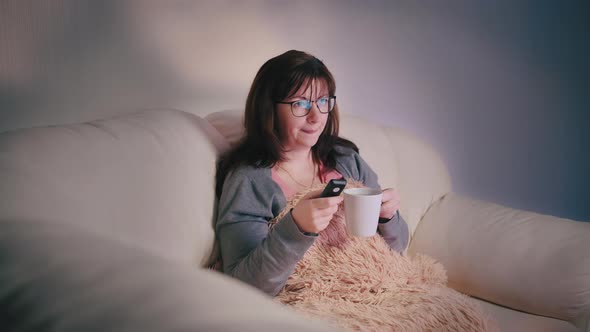 Happy Woman with a Glass of Coffee on the Couch at Night in Front of the TV Switches Channel an