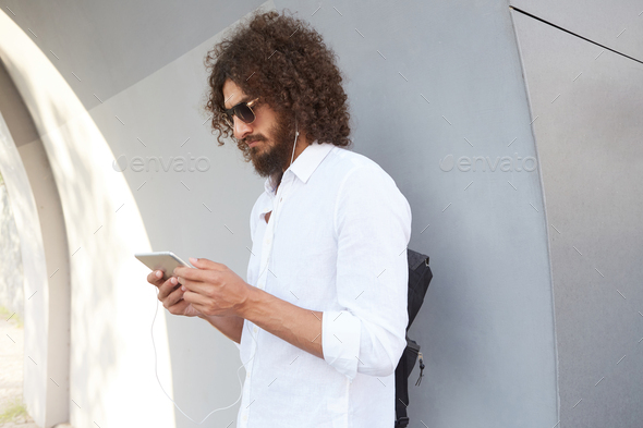 Portrait of handsome dark haired curly man with sunglasses posing over grey  outdoor wall Stock Photo by nakaridore
