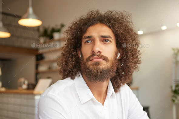 Closeup portrait attractive curly guy with lush beard posing over cafe interior wearing headphones