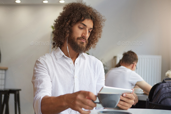 Good looking handsome young bearded man with brown curly hair watching videos on his tablet
