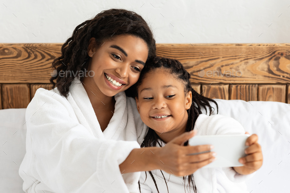 Loving Black Mom Taking Selfie With Daughter, Relaxing On Bed In Bathrobes