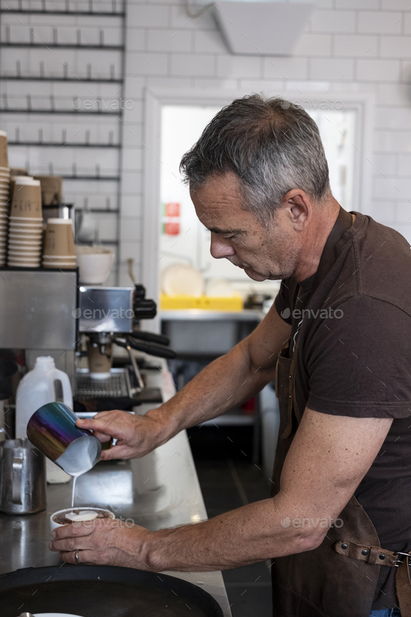 Male barista wearing brown apron, standing at espresso machine, pouring milk.