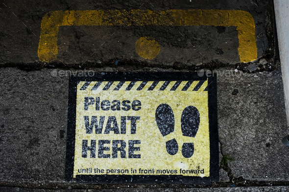 High angle close up of black and yellow \'Please Wait Here\' sign on asphalt ground.