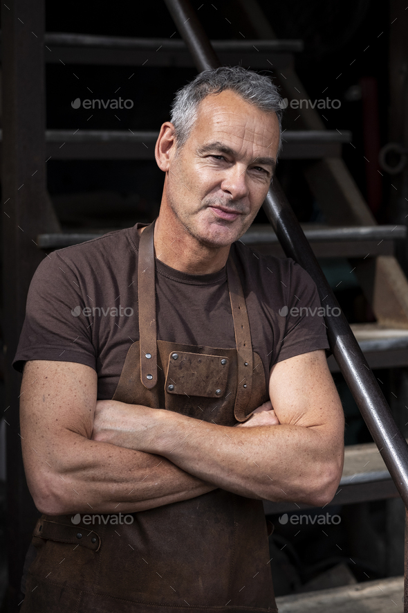 Portrait of male barista with short grey hair, wearing brown apron, arms folded, looking at camera.