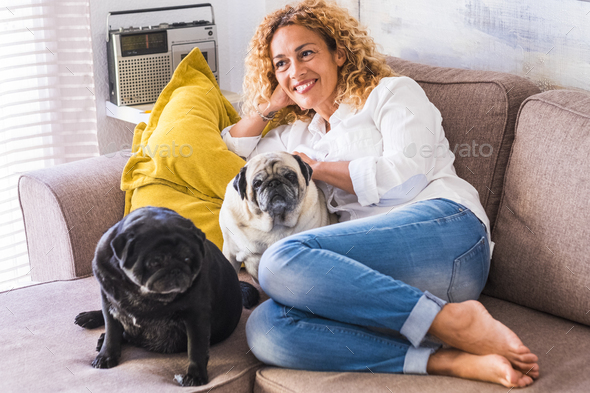 Portrait of cheerful young adult beautiful woman with two dogs pug sitting and relaxing on the sofa