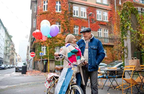Happy senior couple with bicycle and balloons outdoors on street in city.