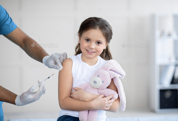 Happy little girl with toy bunny getting coronavirus vaccine injection at hospital. Healthcare and