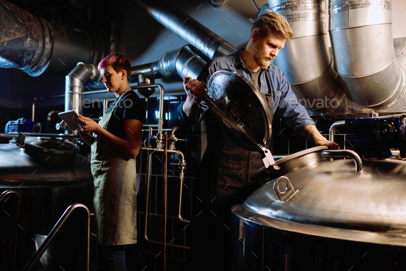Young blond bearded male brewer in workwear opening lid of huge steel tank