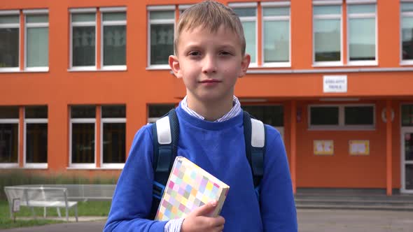 A Young Boy Holds a Book and Looks at the Camera in Front of an Elementary School alt