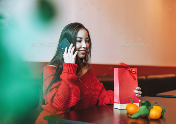 Beautiful smiling young asian woman in red sweater with gift bag using mobile in cafe - Stock Photo - Images