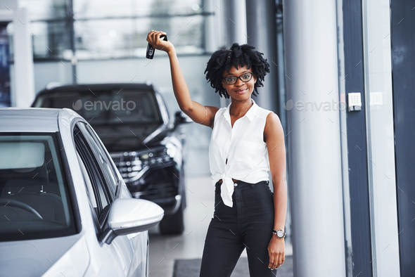 Young african american woman in glasses stands in the car salon near vehicle with keys in hands